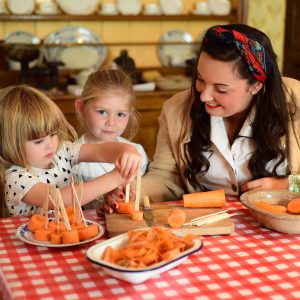 An adult and two children sit at a table with a gingham table cloth pushing lolly sticks into slices of carrot