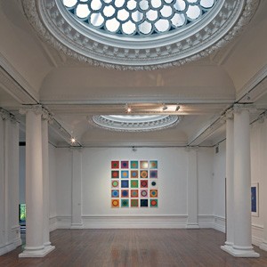 The white interior of the Hatton Gallery featuring two intricately decorated sklylights, columns and a modern graphic artwork on the wall at the end