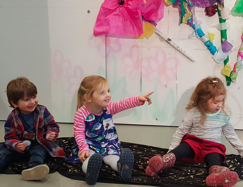 Three young children sit on the floor inside the Northern Gallery of Contemporary Art with colourful craft creations stuck on the wall behind them