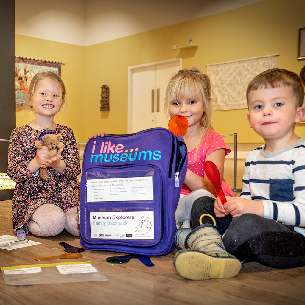 Three children inside a room at Sunderland Museum with an 'I Like Museums' backpack. One is holding a teddy and the others are holding a giant spoon-like object